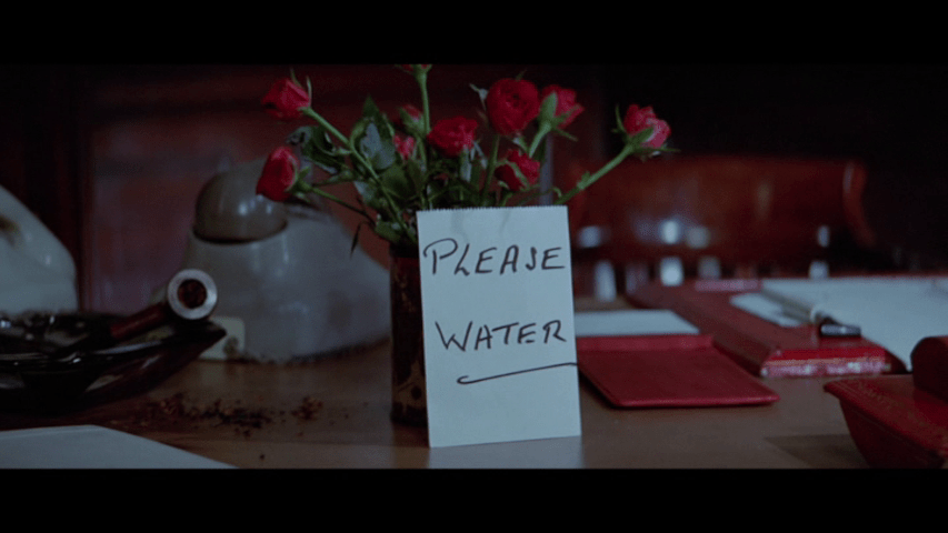 Screengrab from The Tamarind Seed showing a sign reading "Please Water" placed in front of a bouquet of flowers