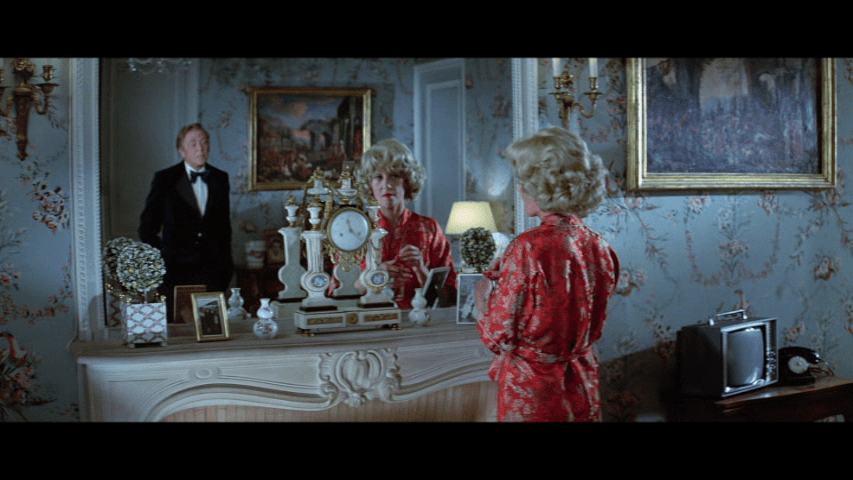 Screengrab from The Tamarind Seed showing a woman and a man standing in front of a mirror with many other things in the frame