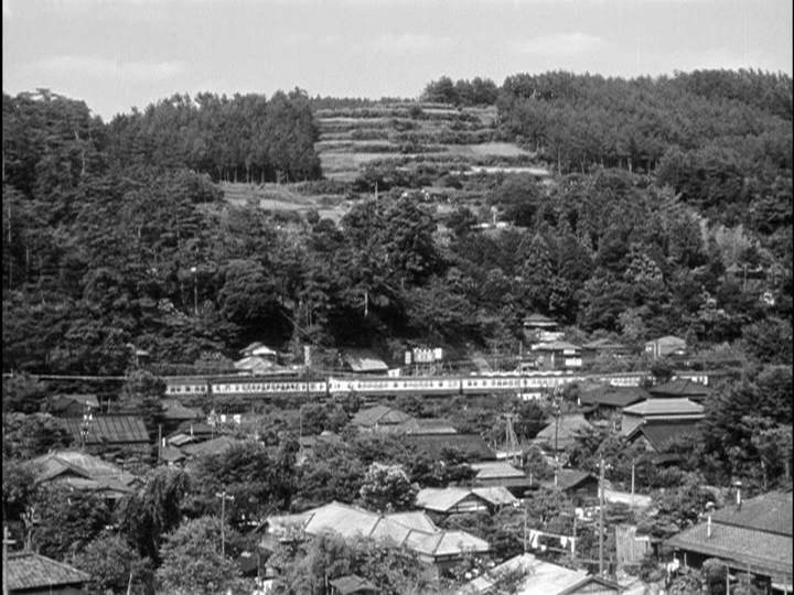 Screengrab from Early Summer showing an exterior with a city in the foreground, forested hills in the background, and a train running through the middle of the frame