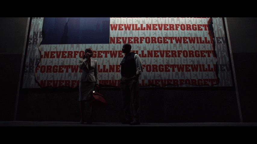 Detective Keith Frazier and Madeleine White talk in long shot in front of an American flag mural where the words "We will never forget" replace the red and whtie stripes