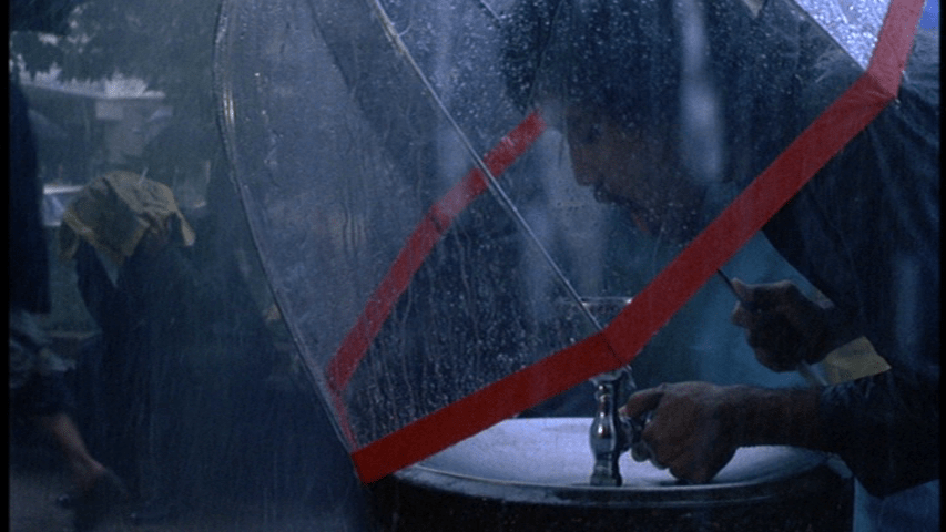 Medium shot of a man with an umbrella drinking from a water fountain during a rain shower