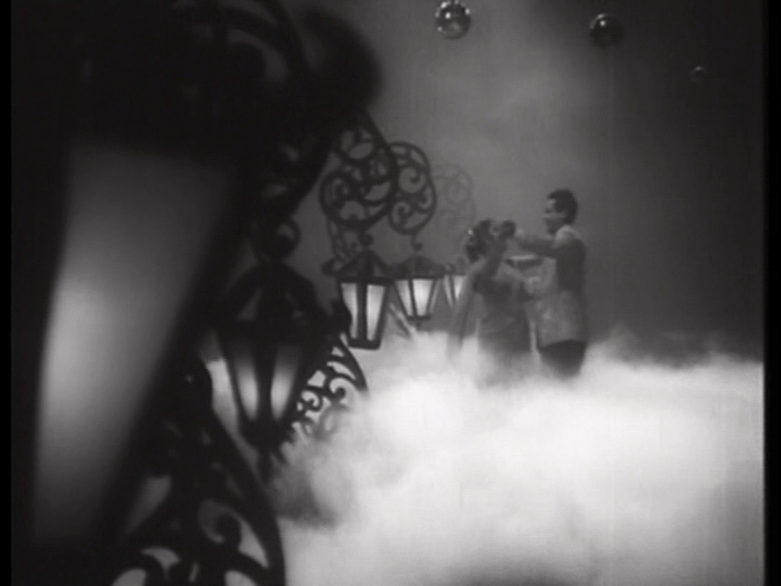 Vijay and Meena waltz on a fog-covered dancefloor next to a row of lamps