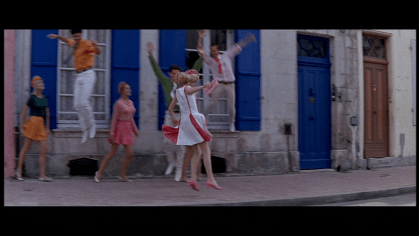 Delphine skips down the street in front of five background dancers