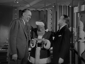 Medium long shot of Kris looking at a check as Misters Gimbel and Macy laugh on either side of him