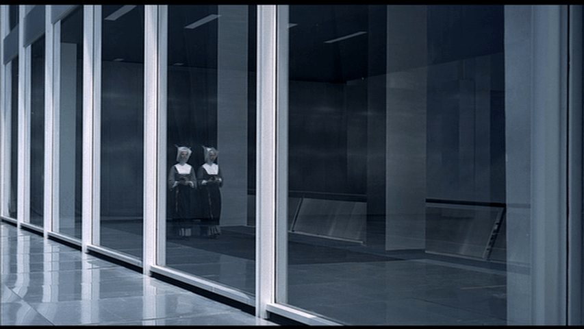 Two nuns walks side-by-side down a corridor which we see through an exterior window