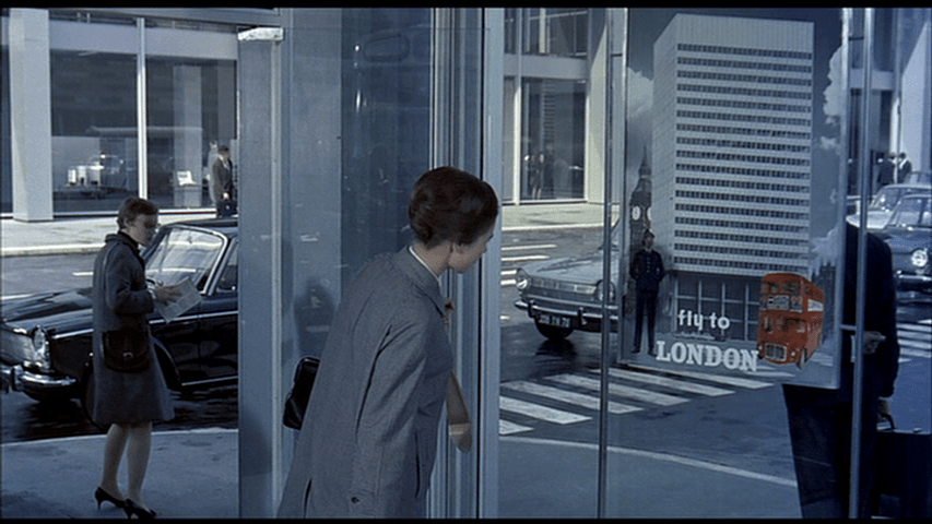 Barbara looks at a poster for London which features a high-rise remarkably like the one she's standing in, only it has a red double-decker bus in front of it