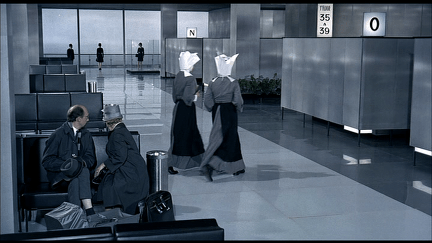 A man and a woman in the foreground look at the two nuns from the previous shot as they pass by