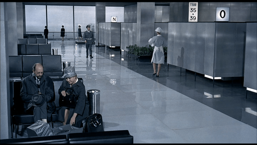 The woman comforts the baby in the carriage we only now see in front of her as a cleaning woman walks away in the background carrying a bundle of towels