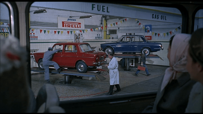 A mechanic leaps onto a hydraulic lift supporting a bright red car next to another one with a blue car on it