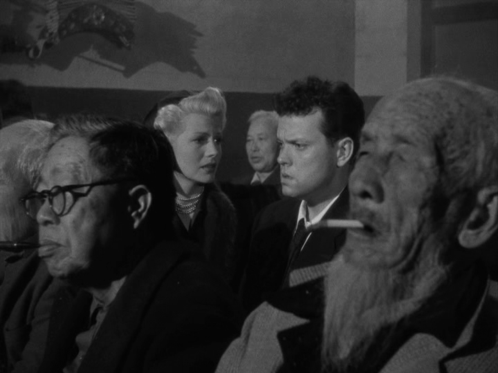 Long shot of Elsa and Michael talk in an audience of theatergoers who could care less