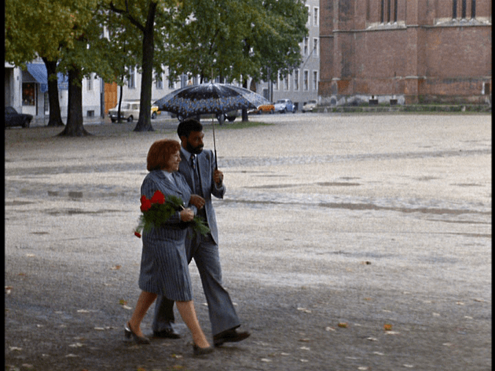 Long shot of Ali and Emmi walking hand in hand: he holds an umbrella, and she has a bouquet of carnations in her arms