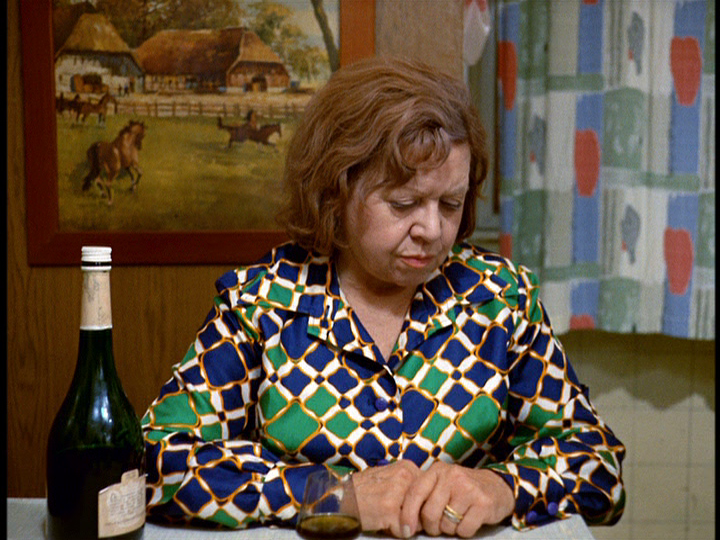 Medium shot of Emmi in the dress and sitting in front of the painting and curtains referenced above