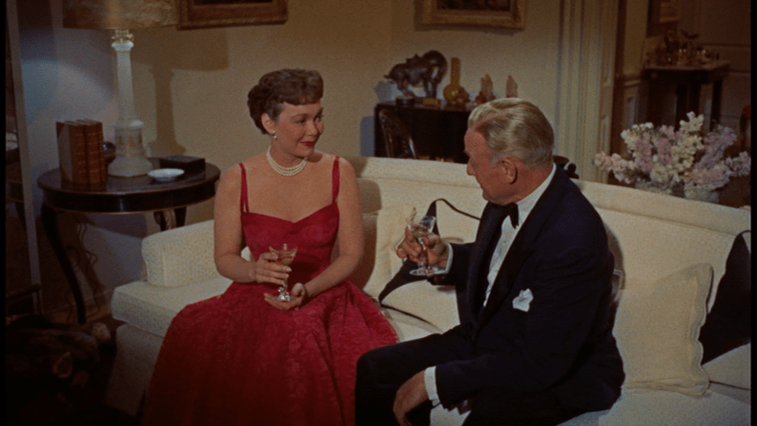 Medium shot of Cary Scott sitting on a couch in a red dress drinking martinis with a man in a tuxedo