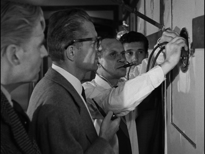 Medium shot of Roger opening a safe as Bob, Paolo, and McKimmie look on