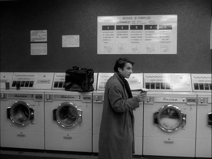 Long shot of Paul facing right in front of a bank of washing machines, gesturing with a cup of coffee