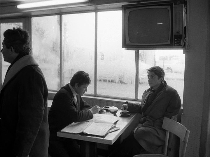 A man asks for directions in the left foreground of the frame while Paul observes him in the right background--Robert is at the same table as him in the middle of the frame