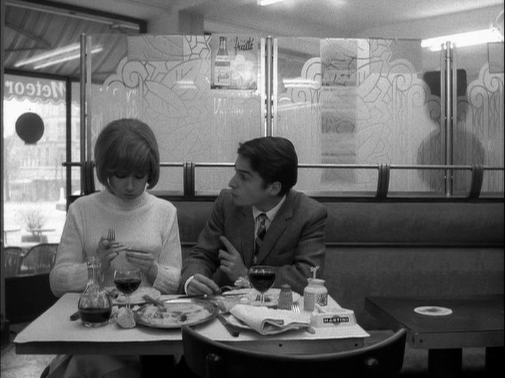 Paul and Elizabeth, facing the camera on the left half of the frame, talk from their seats at a café table covered in food and wine