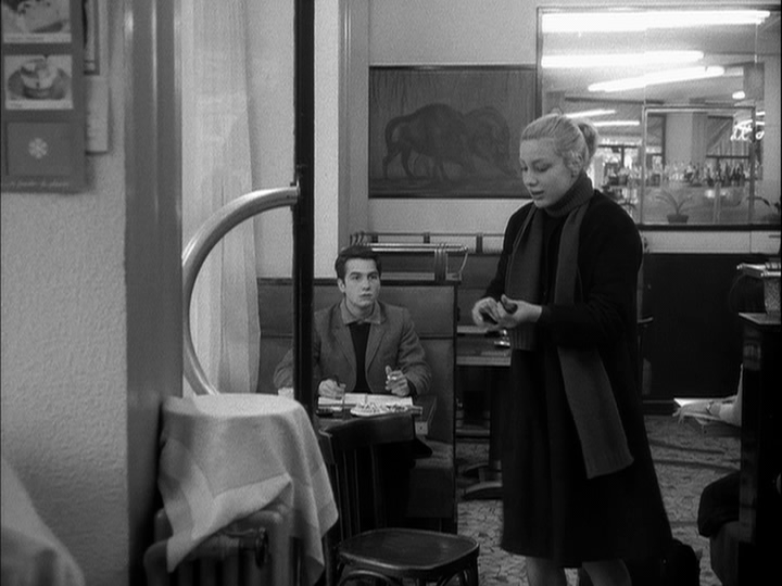 Interior shot of a woman loading a pistol in the foreground as she exists a café while Paul looks on from a table in the background