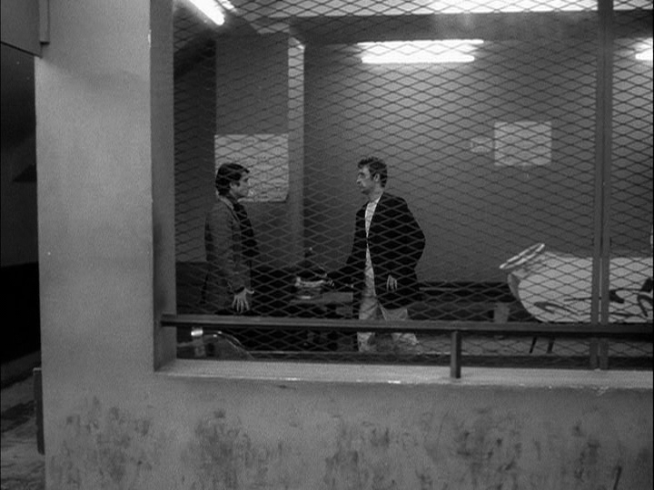 Long shot of a man threatening Paul with a knife, shot through a screened window
