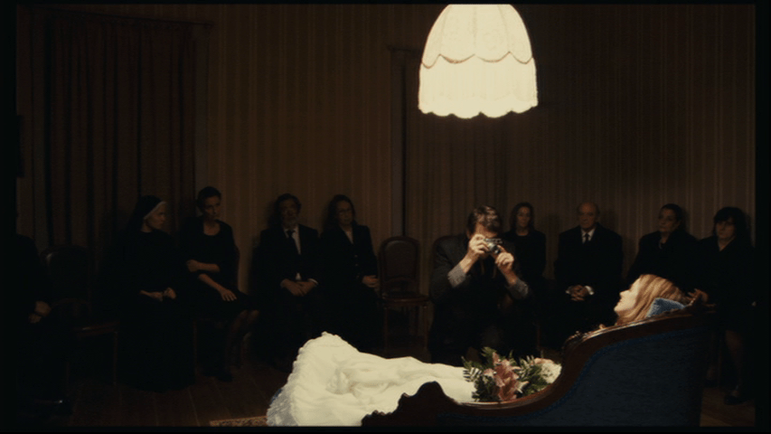 Isaac crouches in the middle right of the frame and takes a picture of Angelica, whose body lies on a fainting couch in the foreground