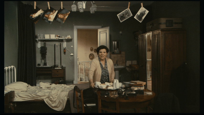 Medium long shot of Madam Justina in Isaac's apartment with a tray of breakfast centered in the frame and looking at the camera
