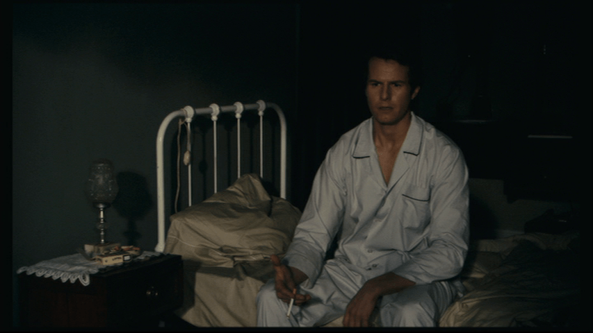 Medium shot of Isaac in night clothes sitting on the edge of his bed smoking a cigarette and talking to himself