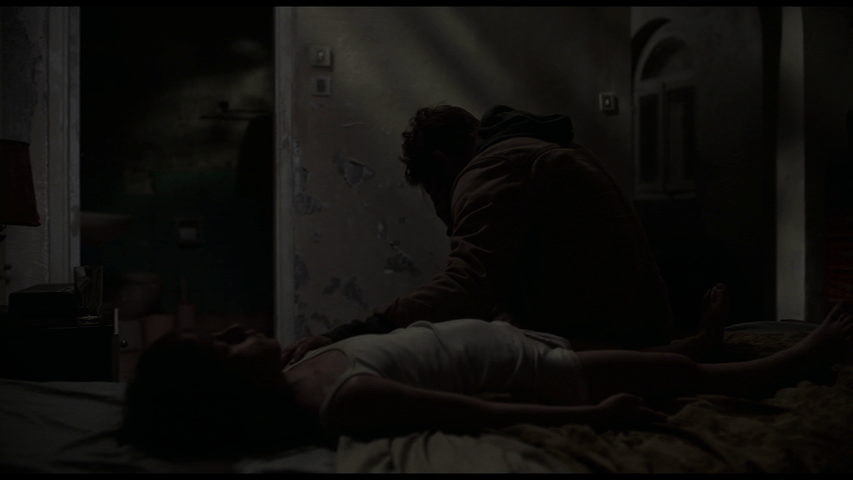 Lang sits on the edge of the bed that Brecken's body is lying on and cries