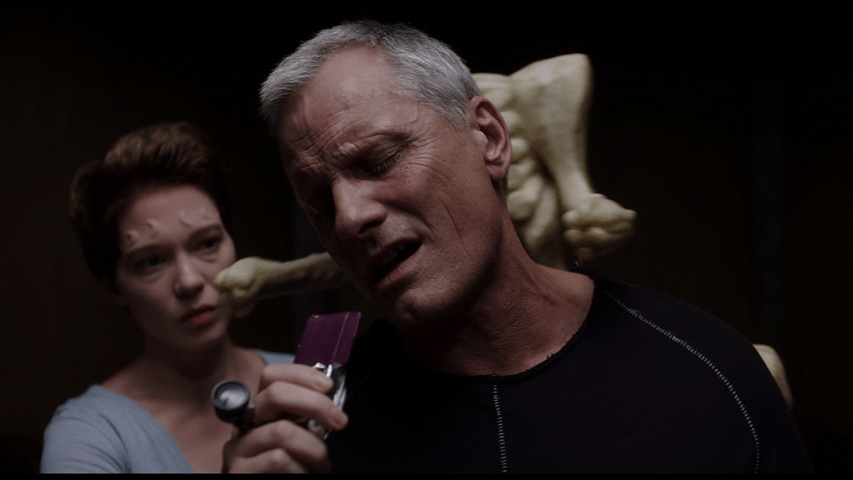 Medium shot of Caprice holding a purple "candy bar" in front of Saul as he sits in his Breakfaster Chair