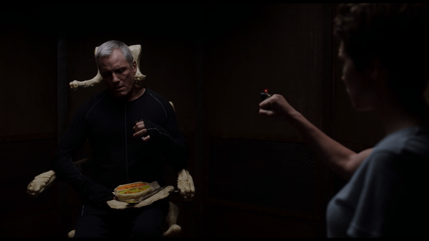 Medium-long shot of Saul in his Breakfaster Chair occupying the left side of the frame as an out-of-focus Caprice films him from the right foreground