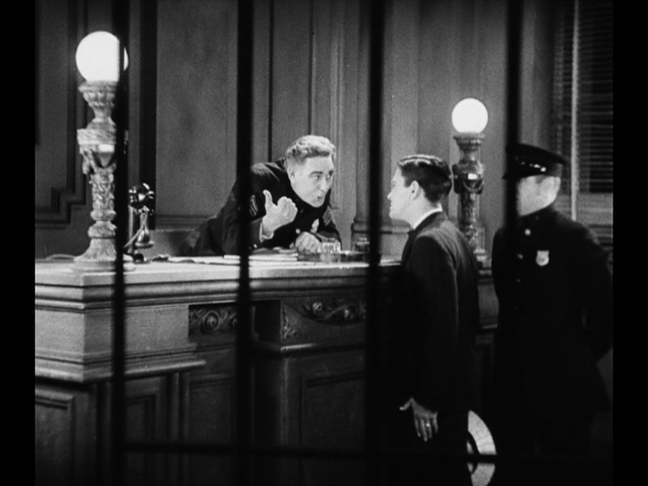 Long shot of Jim arguing his case before two police officers with bars out of focus in the foreground