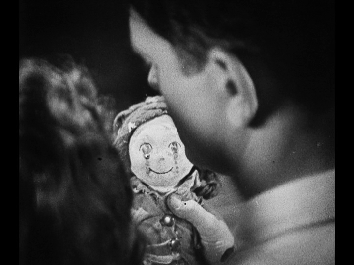 Over-the-shoulder close-up of the doll partially blocked by Jim and Mary's out-of-focus heads in the foreground