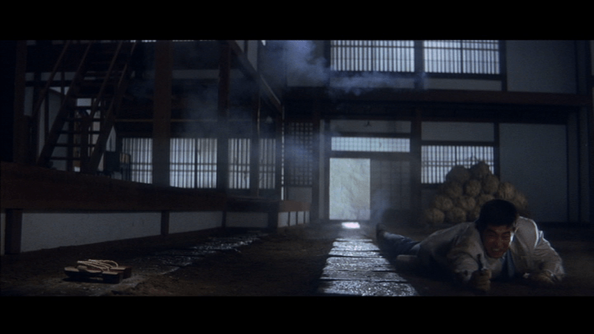 Long shot of Tetsu lying on the ground on the right side of the frame firing his gun toward the camera next to the geta from the previous image, which occupy the left side of the frame