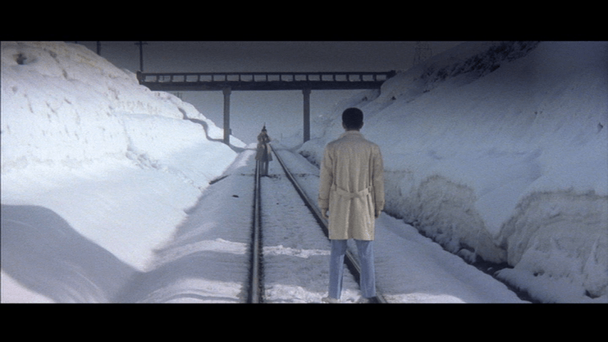 Long shot of Tetsu in the foreground with his back to camera facing off against Tatsu, who aims a gun at him in the middleground