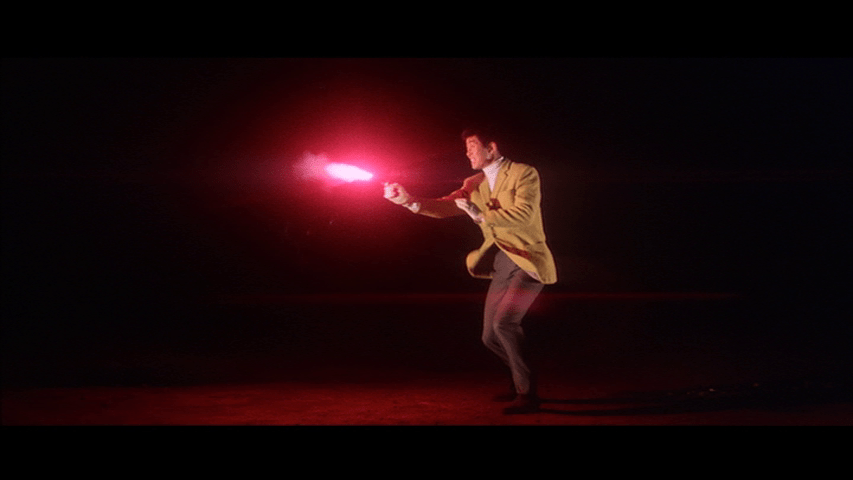 A pink light flashes as Tetsu, clad in a yellow sports coat, shoots his gun at something offscreen facing left against a black background