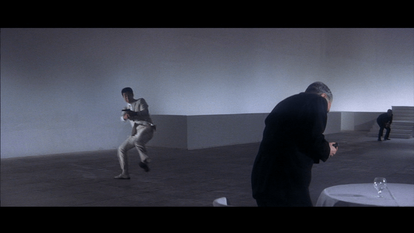 Long shot of Tetsu catching his fun and firing it at Kurata from the background in the left third of the frame as the latter grabs his chest in the right foreground