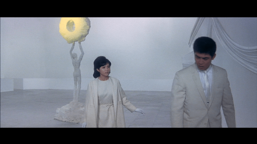 Medium shot of Tetsu on the right side of the frame in an all-white room walking away from Chiharu in the middle of the frame in front of a marble statue holding a doughnut-shaped object lit in yellow