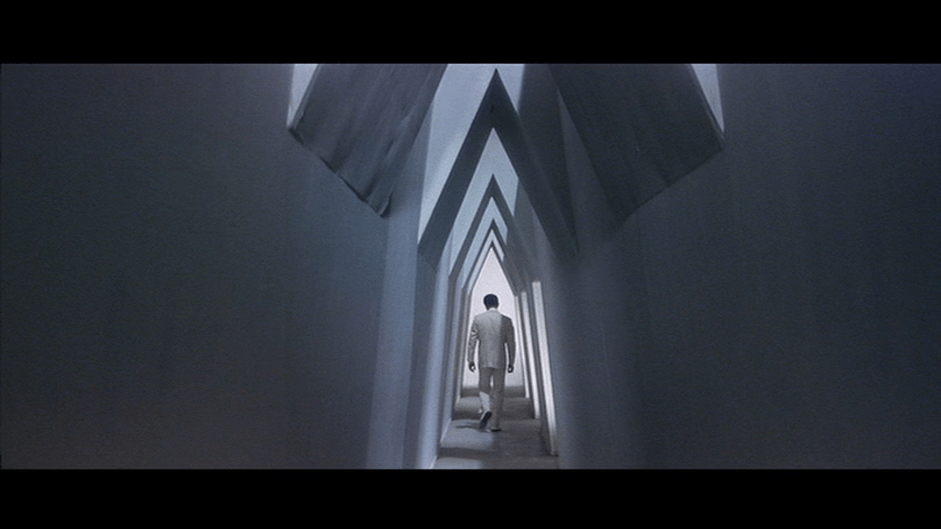 Long shot of Tetsu walking away from the camera in the middle of the frame down a narrow pentagonal hallway