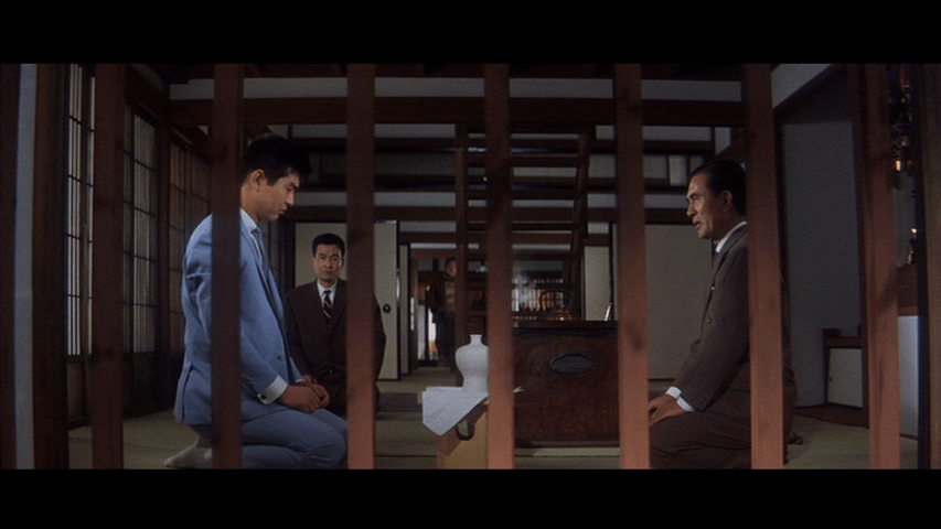 Deep focus long shot composition featuring Tetsu on the left side of kneeling on a tatami mat on the left side of the frame in front of his host who does the same on the right