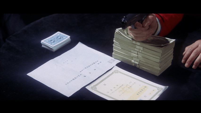 Close up of a hand holding a gun balanced on a stack of money in front of contracts and a deck of cards