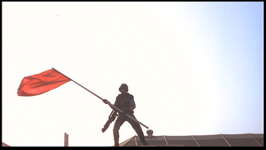 Long shot of Johnny waving a red flag in an image reminiscent of the famous photograph from the Battle of Iwo Jima
