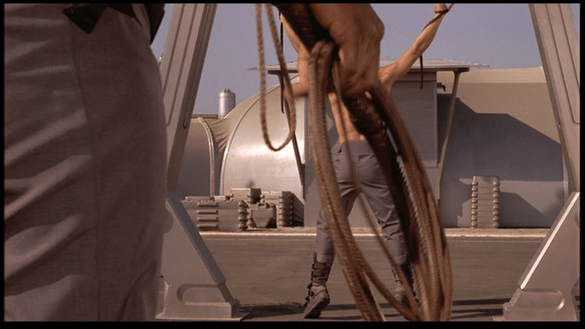 Close-up of a whip being held by an officer which mostly covers Johnny, who faces away from the camera