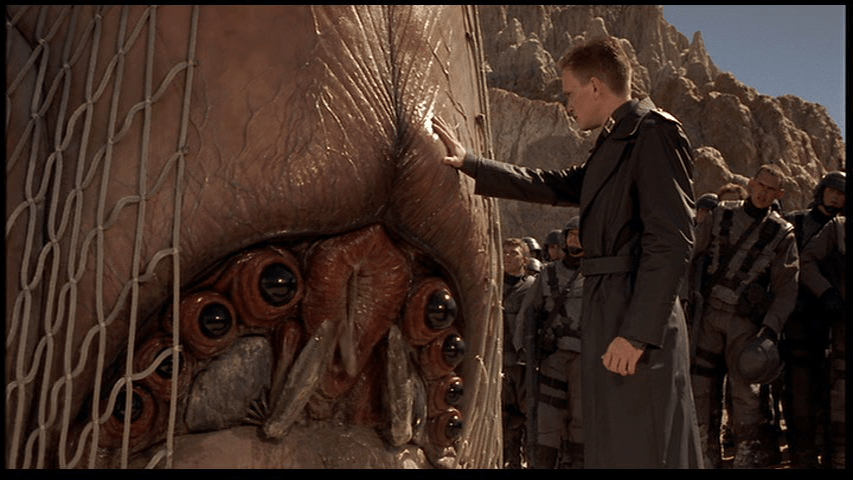 Medium long shot of Carl wearing a uniform very reminiscent of the Third Reich putting his hand on a "Brain Bug" so that he can read its mind