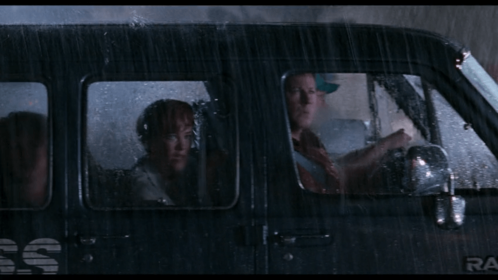 Medium shot of two adults sitting in the front of an airport van looking out a window at a sight that clearly horrifies them