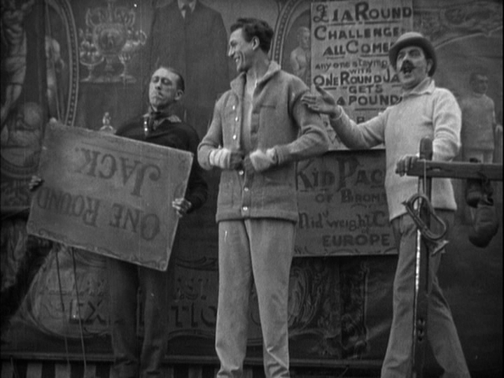 Long shot of Jack Sander in the middle of the frame flanked by his trainer, who holds a sign with his name on it upside down, to his right and Harry Terry's The Showman to his left
