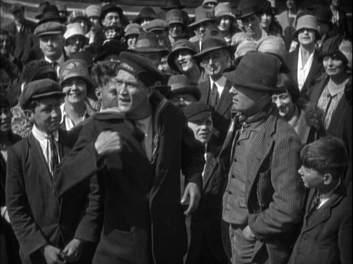 Long shot of a big sailor pointing at his chest in the midst of a crowd