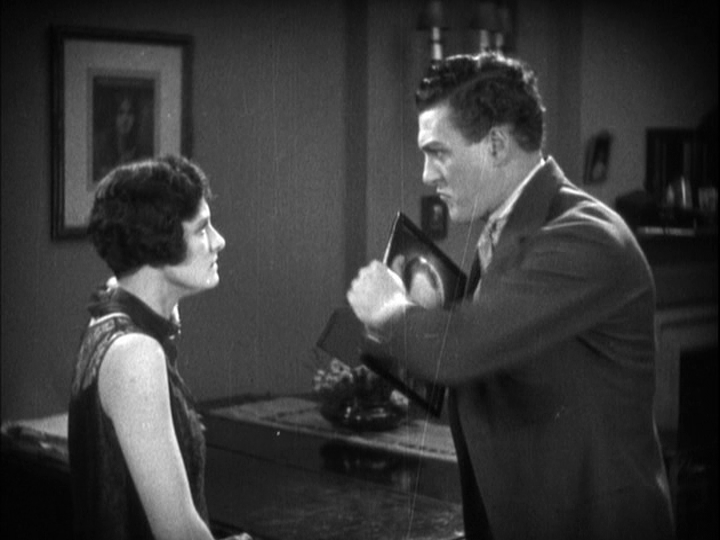 Medium shot of Sander preparing to throw a framed photo of Corby on the right side of the frame while Mabel glares at him from the left