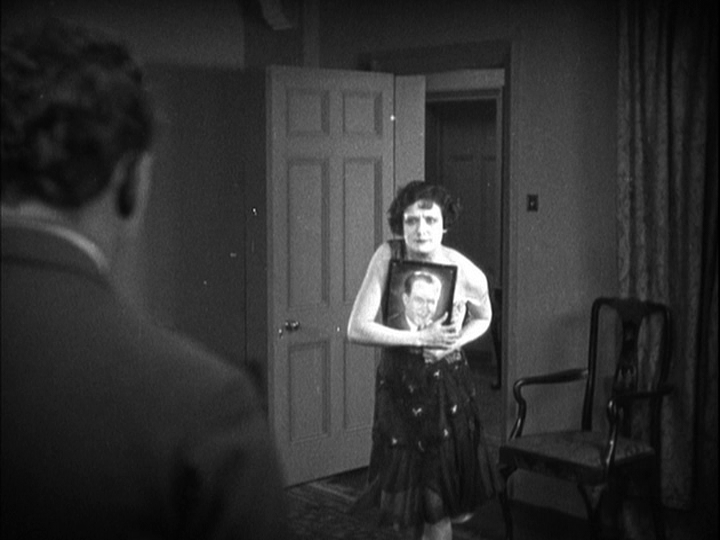 Medium shot of Mabel clutching Corby's photo to her exposed chest as she backs away from Sander, who occupies the left third of the frame in the foreground