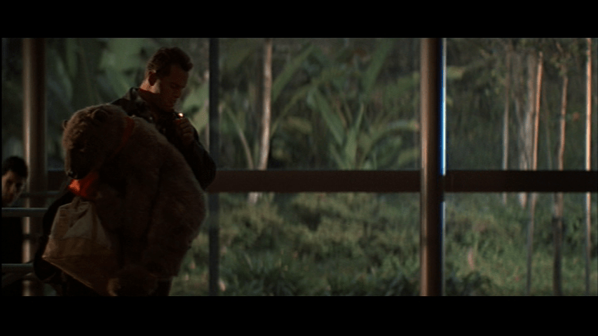 Medium long shot of John McClane holding a large stuffed bear and lighting a cigarette in the left third of the frame
