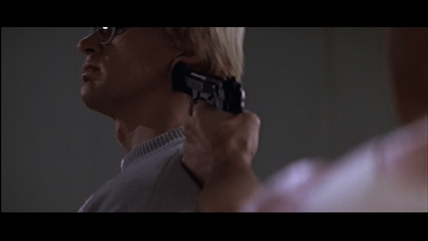 Close-up of McClane out of focus in the right foreground holding a gun on Tony, who is now in focus in the left background