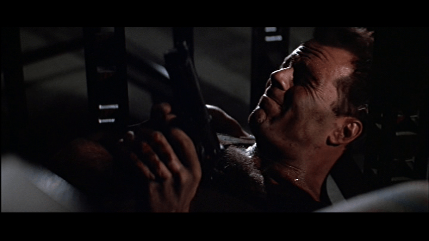 Close-up of McClane under the table aiming his guns upward at Marco